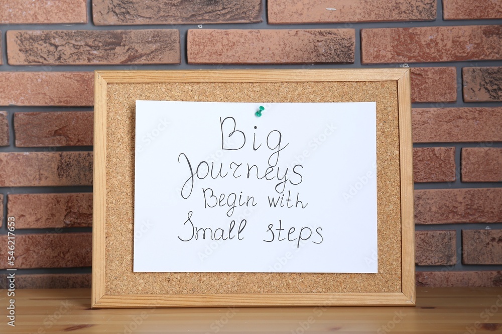 Obraz premium Corkboard and phrase Big Journeys Begin With Small Steps on table against brick wall. Motivational quote