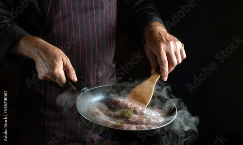 A professional chef fries hot dog sausages in a frying pan. Close-up of a cook hands with a hot pan in the kitchen. Free space for advertising on a black background