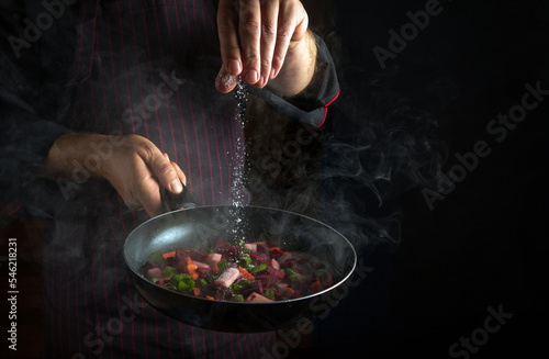 Fototapeta Naklejka Na Ścianę i Meble -  A professional chef adds salt to a steaming hot pan. Molecular cuisine menu idea for a hotel with advertising space on a black background