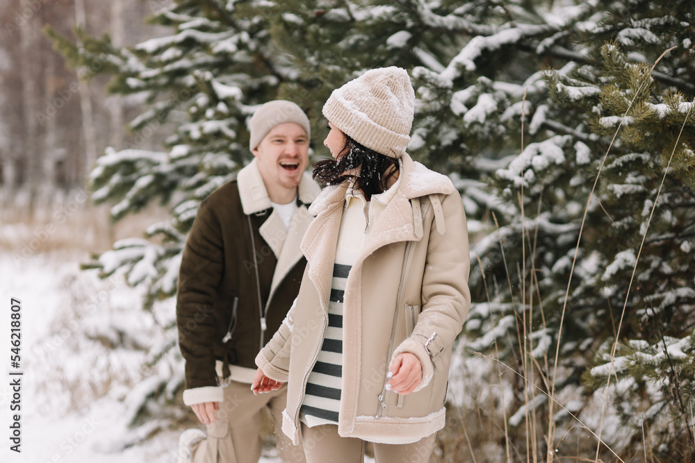 A couple in love spend time together walking in a snowy forest among trees having fun and laughing in nature on vacation. A young man and a woman in warm clothes travel outdoors. Selective focus