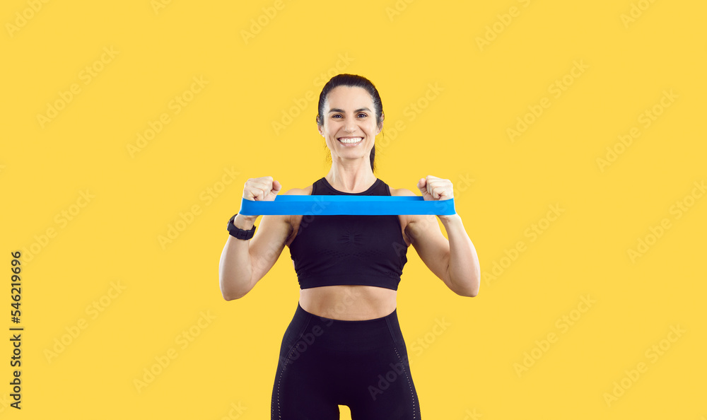 Cheerful sports woman on yellow background with her hands stretches rubber elastic band for training. Portrait of beautiful smiling woman with slender body who is wearing black sportswear set. Cheerful sports woman on yellow background with her hands stretches rubber elastic band for training. Portrait of beautiful smiling woman with slender body who is wearing black sportswear set.