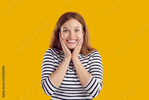 Portrait of beautiful young woman isolated on yellow background holding hands on face, smiling and looking camera like at wonderful surprise with happy, excited face expression
