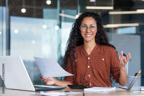 Portrait of happy and successful hispanic woman, businesswoman smiling and looking at camera holding contracts and invoices, working inside office with laptop on paper work.