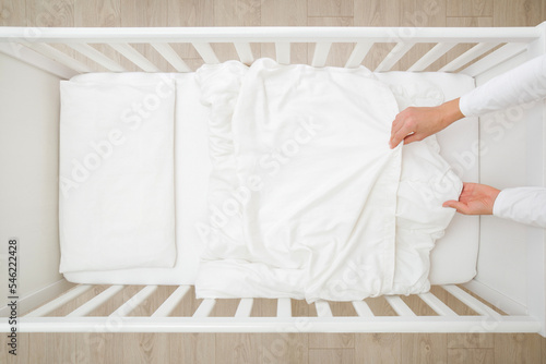 Young adult mother hands changing white blanket cover in baby crib. Regular bed linen change. Closeup. Top down view.