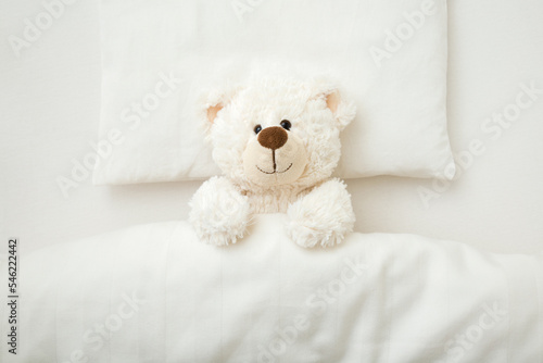 Adorable smiling white teddy bear lying down on pillow and sheet under blanket in baby crib at home. Closeup. Top down view.