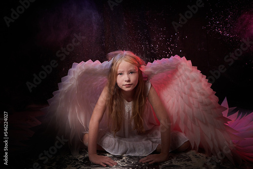 A girl in a white shirt, wings and with long hair, looking like a angel. A young model posing at a photo shoot with smoke, fog, flour, dust. A little fairy girl on a dark black background photo