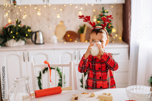 A charming boy in plaid pajamas, drinking milk in a kitchen decorated for Christmas. The concept of comfort and celebration