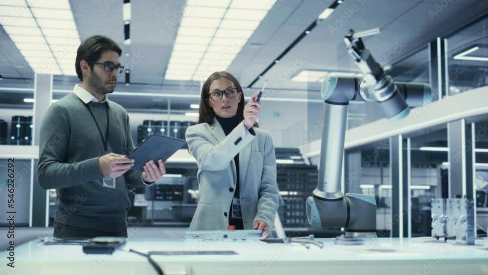 Robotics Engineer and Machinery Operator Collaborate on a Robot Arm Project at a Factory, Using a Tablet Computer to Operate the Robot Arm in Scientific Technology Lab. Robotics Engineer and Machinery Operator Collaborate on a Robot Arm Project at a Factory, Using a Tablet Computer to Operate the Robot Arm in Scientific Technology Lab.