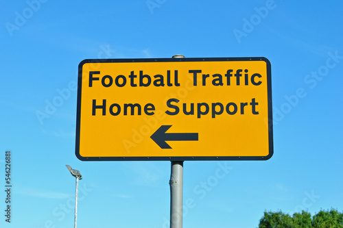 Close Up of Yellow Road Sign 'Football Traffic Home Support ' on Pole against Blue Sky