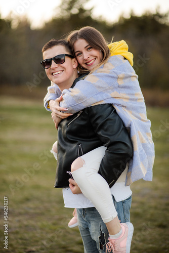 a young couple, a girl on the back of a guy, he is in a leather jacket and sunglasses, she is dressed casually, they are smiling and happy