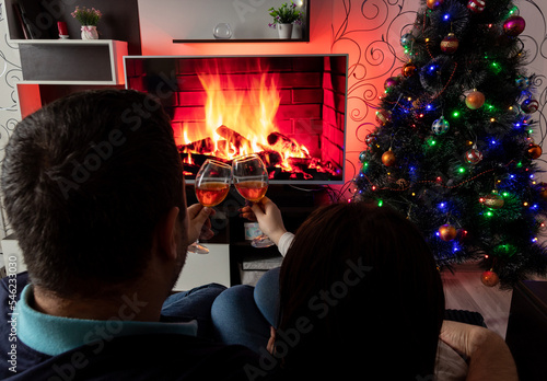 Couple is sitting on sofa, touching glasses of wine, in front of TV, which depicts fireplace. Selective focus. Images for articles about leisure, winter, family
