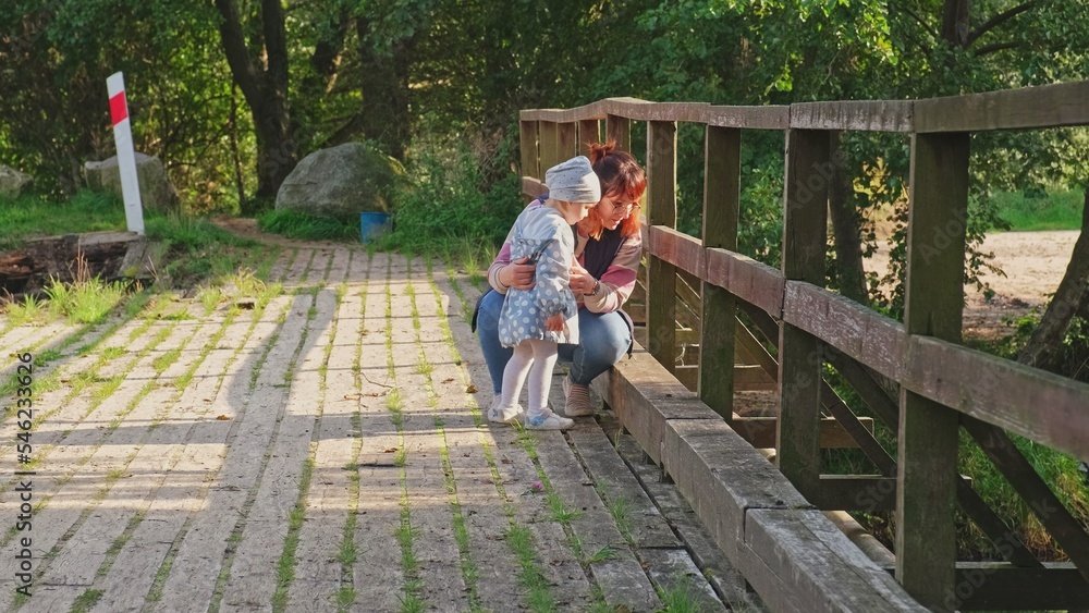 Fototapeta premium Caucasian Woman Standing on Wooden Bridge with Her Baby Toddler Daughter Looking Down at River Water
