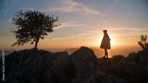 A young woman stands at sunset in the mountains. The silhouette of a girl against the background of the sunset sky and the sea on the top of the mountain. The girl stands on the viewpoint and enjoys t