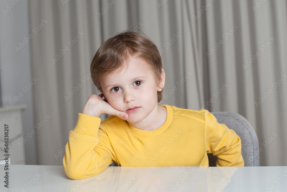 Pensive child boy with brown hair in yellow sweater sitting by the table and looking to the camera on beige curtain background Pensive child boy with brown hair in yellow sweater sitting by the table and looking to the camera on beige curtain background