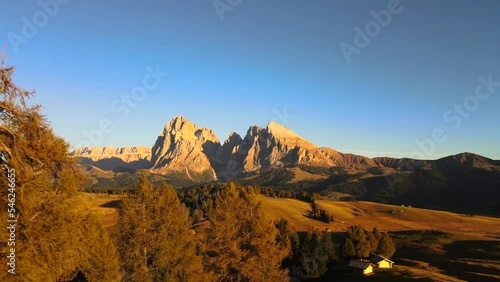 Mountains, forest and grass fields with wooden cabins filmed at Alpe di Siusi in Alps, Italian Dolomites filmed in vibrant colors at sunset. Filmed with a drone flying backwards over trees, wide view photo