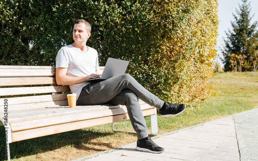 Remote work, freelance concept. Young man sitting with laptop and cup of coffee on a park bench looking away on a sunny day outdoors Remote work, freelance concept. Young man sitting with laptop and cup of coffee on a park bench looking away on a sunny day outdoors