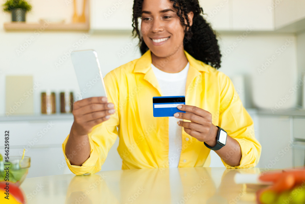 Happy african american lady with smartphone and credit card, shopping online in kitchen, ordering groceries remotely Happy african american lady with smartphone and credit card, shopping online in kitchen, ordering groceries remotely
