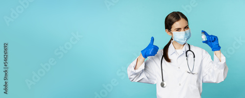 Portrait of smiling doctor, medical personel in face medical mask and rubber gloves, showing thumbs up and coronavirus, omicron vaccine, standing over blue background