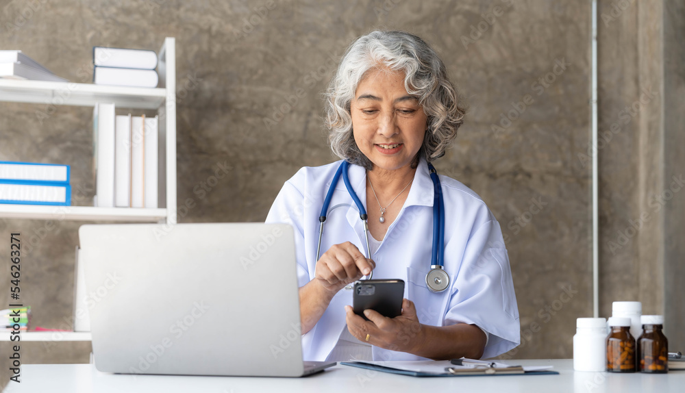 doctor senior grey-haired woman in white medical coat is using a smartphone and smiling while sit use laptop at office doctor senior grey-haired woman in white medical coat is using a smartphone and smiling while sit use laptop at office