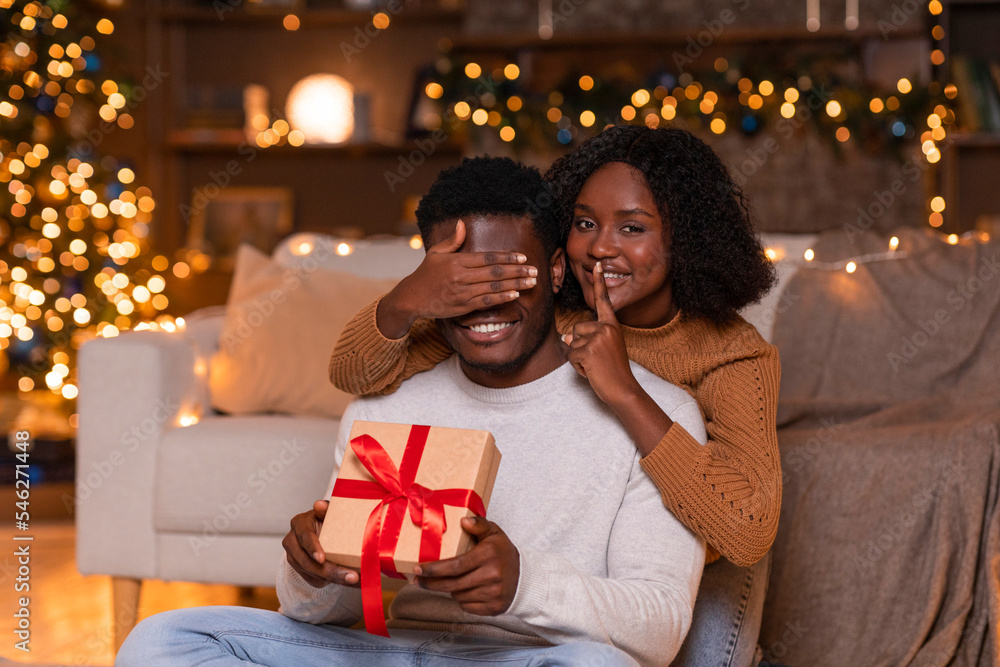 Smiling young african american wife closes eyes to husband, shows silence sign and gives gift box Smiling young african american wife closes eyes to husband, shows silence sign and gives gift box