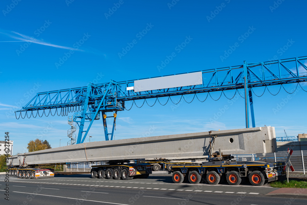 Overhead crane and trailer with long concrete beams against the blue sky. On the side of the road.