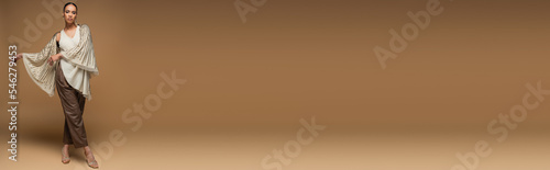 Bild auf Leinwand full length of stylish african american woman in golden shawl and leather pants posing on beige, banner
