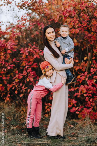 Photography  a portrait of a beautiful brunette girl with a small child  a boy  a son  and a smiling girl  a daughter  in nature in autumn  against the backdrop of a tree with red leaves.