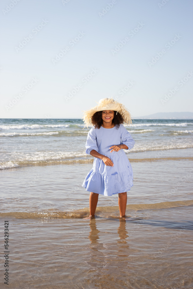 Delighted little girl in hat and dress on beach. African American child on beach on summer day, standing in waves. Childhood, vacation, happiness concept