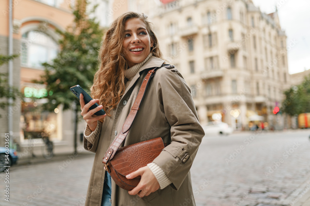 Smiling curly woman wearing warm coat walking down the street and using her phone