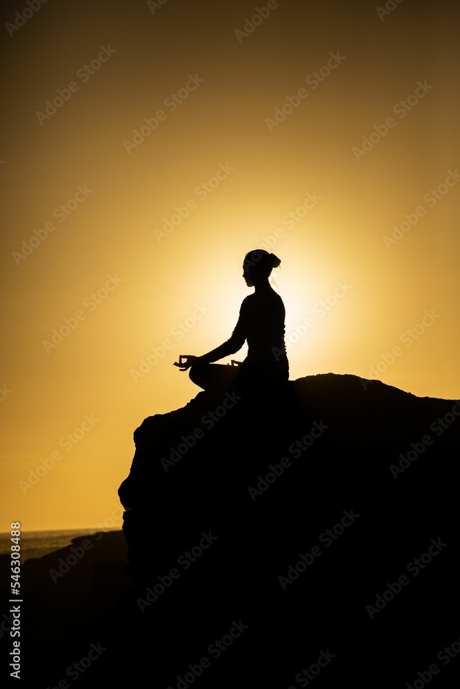 Vertical shot of female silhouette meditating on the edge of the mountain at sunset