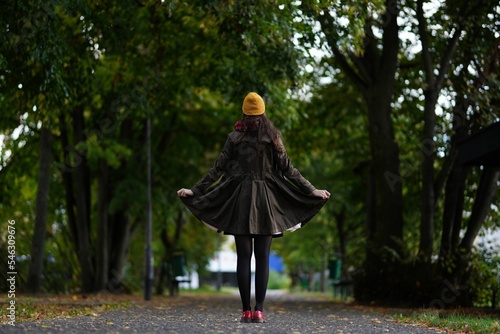 Female standing in olive coat, yellow hat and red shoes on an alley with green trees during autumn