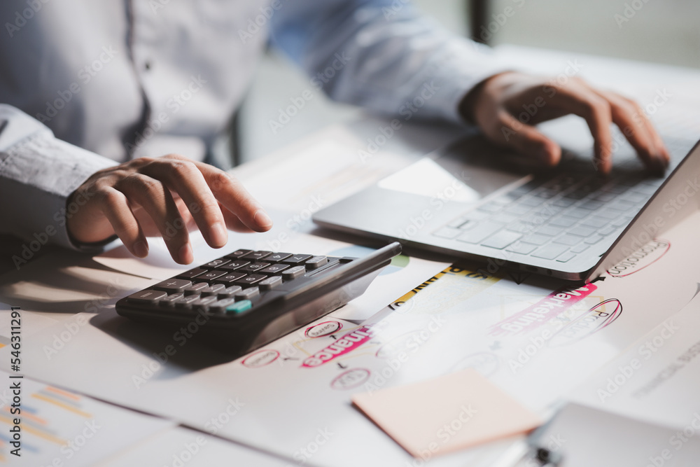 Fototapeta premium Businessman is using a calculator to calculate company financial figures from earnings papers, a businessman sitting in his office where the company financial chart is placed.