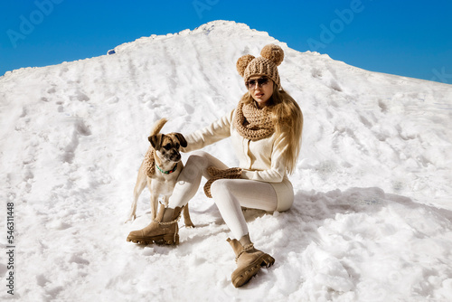 Beautiful stylish woman playing with her dog in the snow in winter scenery in the mountains. Winter vacation.