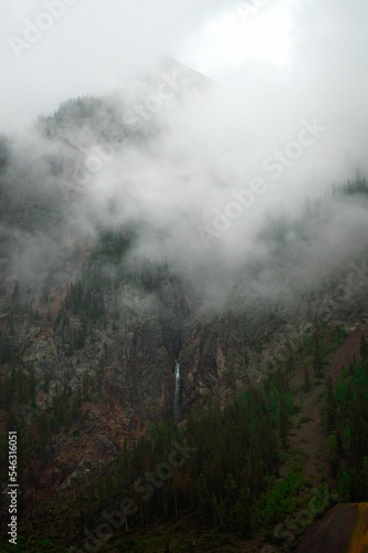 Vertical low-angle of a small waterfall on the large mountainside, misty and lush greenery around