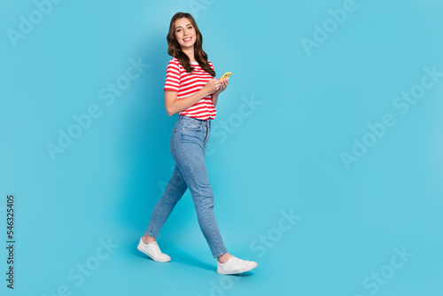 Full length photo of funny sweet lady wear striped t-shirt walking chatting modern device empty space isolated blue color background