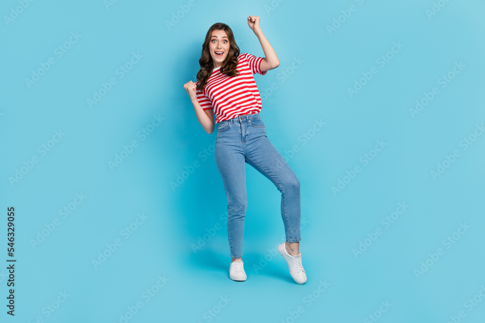 Full length photo of lucky excited lady wear striped t-shirt smiling rising fists isolated blue color background Full length photo of lucky excited lady wear striped t-shirt smiling rising fists isolated blue color background