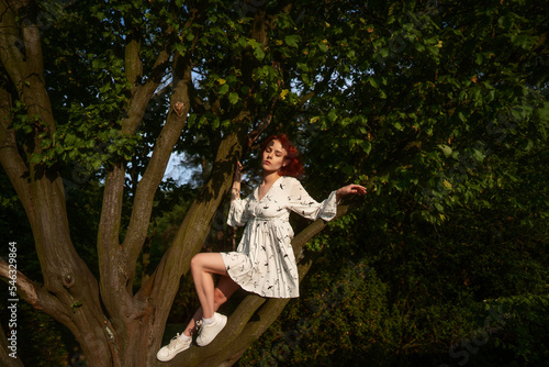 Slim lovely girl relaxes in the park before sundown. Fashion photo as poster or wallpaper. Attractive red haired woman in a white dress sits on the trees branch and poses in the light of the sunset.