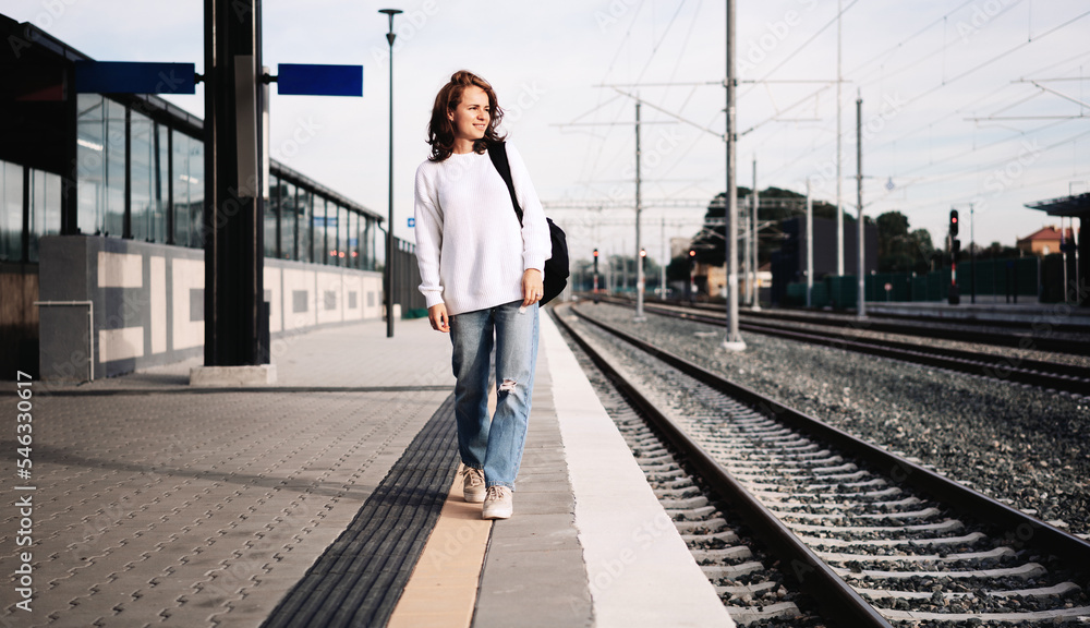 Woman wearing casual white clothes walking on railroad station platform with backpack.