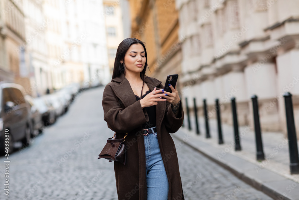 trendy brunette woman with crossbody using mobile phone on blurred street in prague. trendy brunette woman with crossbody using mobile phone on blurred street in prague.