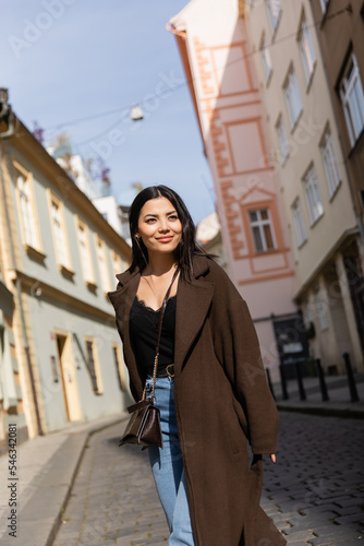 Cheerful woman in brown coat looking away on street with sunlight in Prague. © LIGHTFIELD STUDIOS