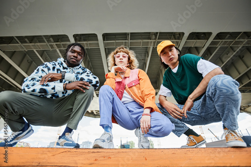 Group of three young people wearing street style clothes outdoors while sitting on stairs in urban area and looking at camera photo