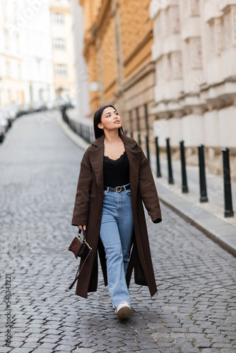 full length of woman in stylish coat an jeans walking in prague and looking away.