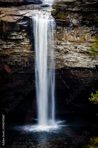 High waterfall cascading down cliff along rock face into pool of fresh water near cave  vertical frame