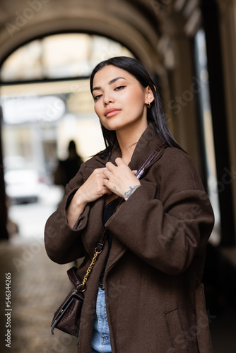 young brunette woman in trendy brown coat with crossbody on blurred background in prague. photo