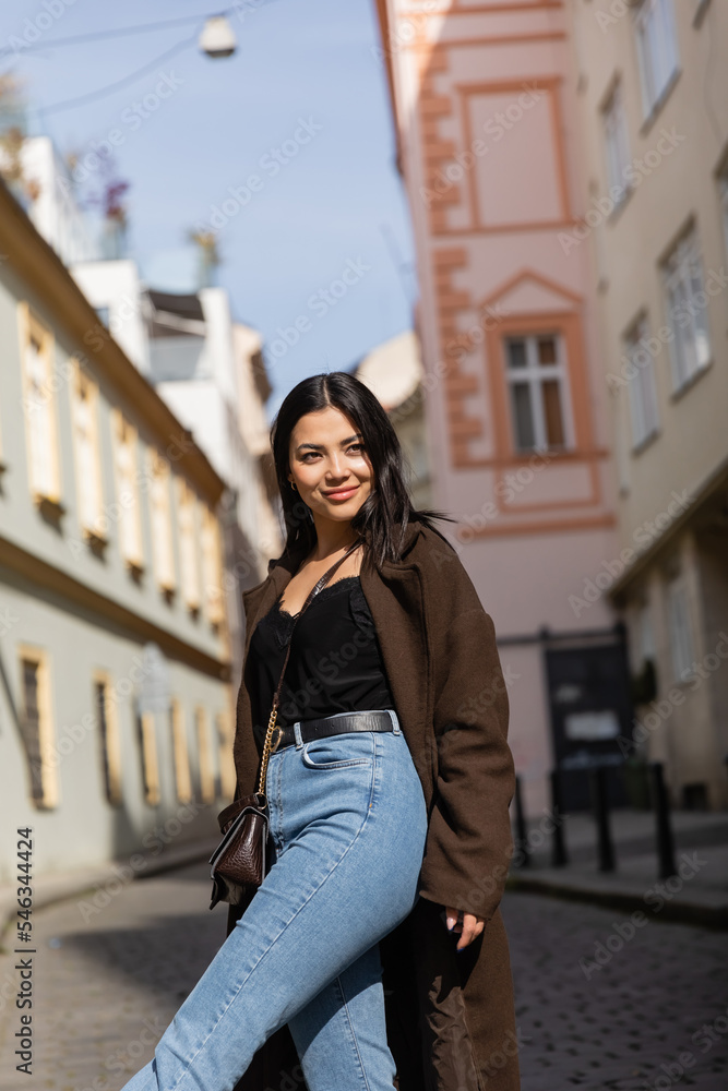 Happy brunette woman in coat and jeans looking away on street in Prague.