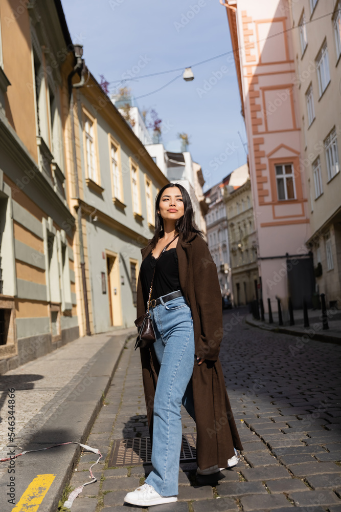 Full length of stylish woman in coat walking on road between buildings on street in Prague.