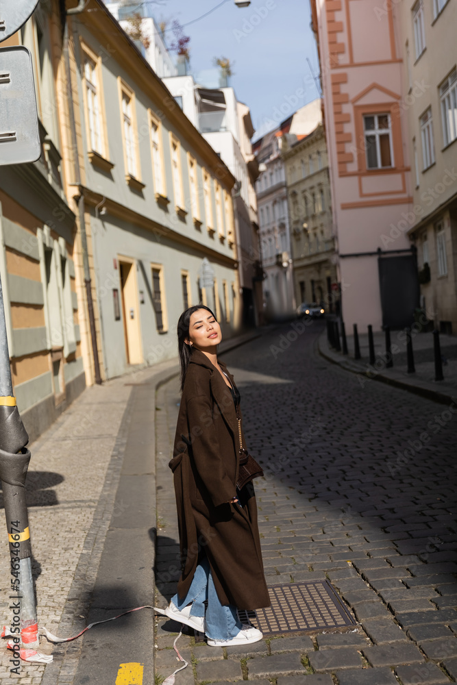 Brunette woman in coat walking on blurred urban street in Prague.
