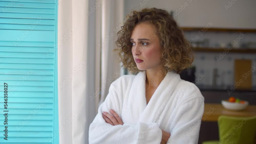 Sad worried young woman in bathrobe standing near window at home. Realtime Sad worried young woman in bathrobe standing near window at home. Realtime
