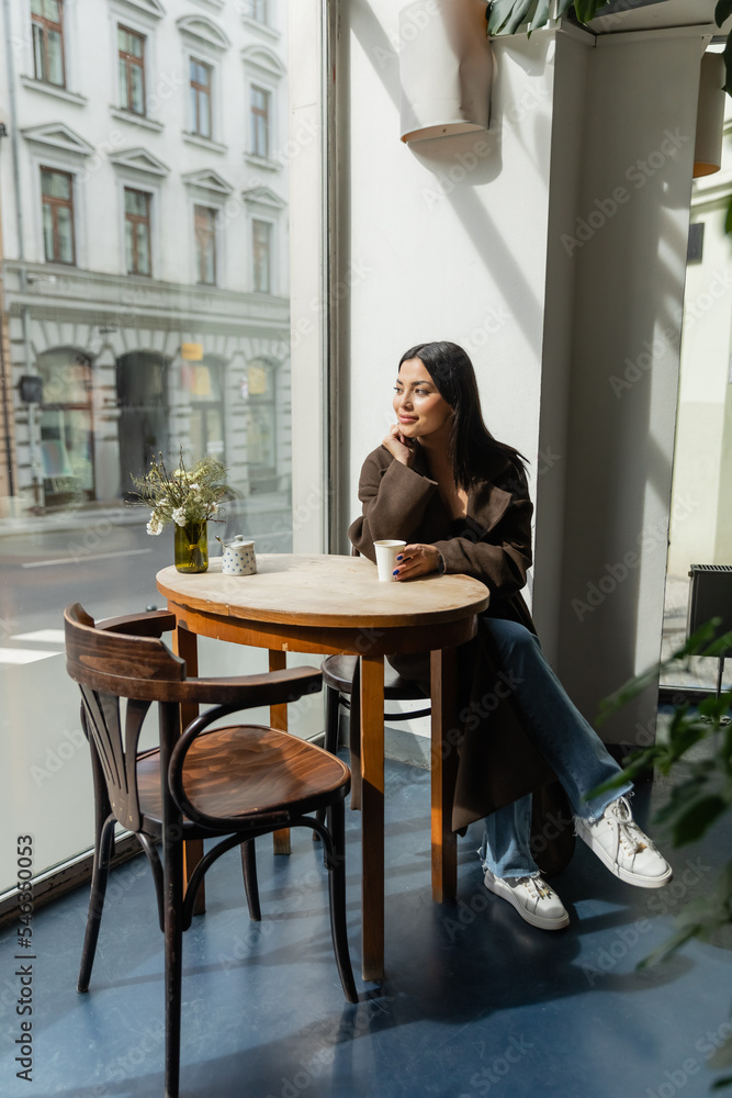 young woman in autumn coat sitting in cafe near window and looking at street in prague.
