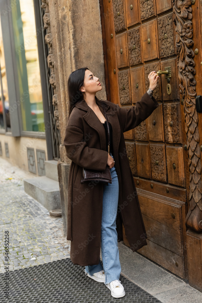 full length of brunette woman in stylish coat with crossbody opening carved wooden door of building in prague.